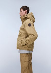 Νέος άνδρας με μπεζ μπουφάν με κουκούλα και επιδιορθωτική ρεβέρ στο μανίκι, καθώς και ταιριαστόι παντελόνια cargo, στέκεται προφίλ με τα χέρια στις τσέπες.
