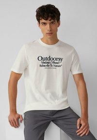 Witte t-shirt met korte mouwen van katoen, met een donkerblauwe grafische tekstontwerp dat "Outdoorsy Feeling Ahead! Abonneer je op de Natuur" leest.
