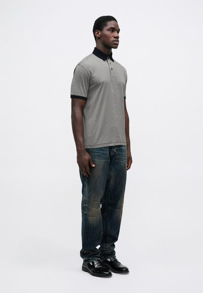 Pilka ir juoda dryžuota polo marškinėliai su juodu apykakle, derinami su išblukusiais mėlynais džinsais ir juodais batais. Modelis stovi tiesiai prieš paprastą foną.