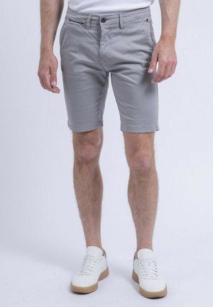 Homme portant un short gris clair longueur genou, des baskets blanches et une chemise blanche sur un fond uni.