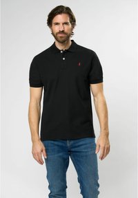 Black polo shirt in cotton blend with short sleeves, featuring a two-button placket and a small red logo on the chest. Worn with blue jeans.