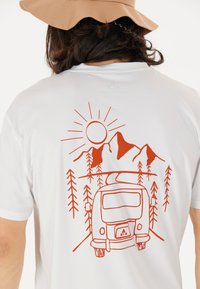 Vit t-shirt med en röd grafisk design av en vintage husvagn, berg, sol och träd på baksidan. Korta ärmar, avslappnad passform.
