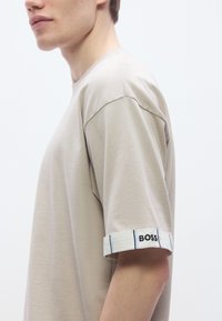 Beiges Baumwoll-T-Shirt mit kurzen Ärmeln. Die Bündchen sind mit einem weißen Band mit blauen Streifen und dem schwarzen Stickschriftzug „BOSS“ verziert.