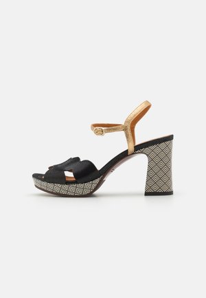 Schwarze und goldene Absatzsandalen mit strukturierten Riemen, offener Zehenpartie und geometrisch gemusterter Plattform. Großer Absatz mit kontrastierenden Designdetails.