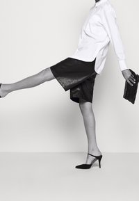 Camicia bianca a maniche lunghe, pantaloni corti in pelle nera e décolleté neri a punta. Modella in posa con una gamba sollevata, mentre tiene una pochette nera con texture.