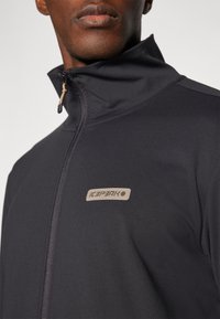 Giacca nera con zip e collare alto, con una patch del logo beige sul davanti. Texture liscia con un design aderente.