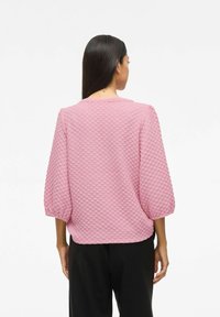 Kvinna med långt mörkt hår iklädd en texturerad rosa topp med trekvartslånga ärmar och svarta byxor, sedd bakifrån mot en vit bakgrund.