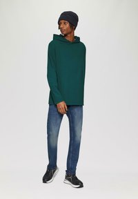 Zielona bluza z długim rękawem i kapturem, zestawiona z jasnoniebieskimi dżinsami. Czarne buty sportowe. Prosty, casualowy design bez widocznych wzorów.