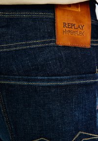 Lähikuva tummansinisten farkkujen takataskusta, jossa on vaaleanbeige tikkaus ja ruskea nahkalappu, johon on kohokuvioitu "REPLAY HYPERFLEX."