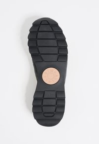 Black rubber sole with textured tread pattern, featuring a circular logo in tan at the center. Upper section shows a gray textile finish.