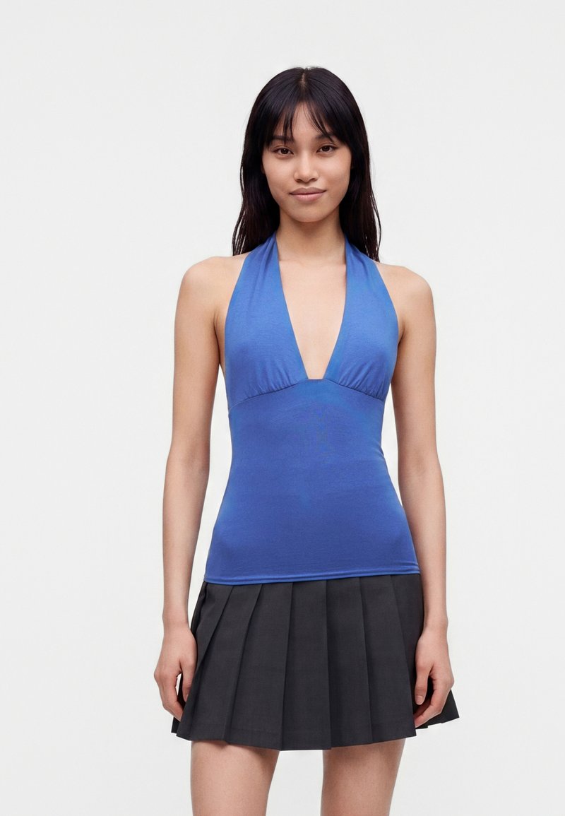 Femme aux longs cheveux foncés portant un haut bleu à bretelles croisées et une jupe noire plissée, debout devant un fond clair uni.