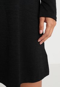 Abito nero lavorato a maglia con maniche lunghe, caratterizzato da una consistenza morbida e una vestibilità rilassata, tenuto da una mano vicino all'orlo per evidenziare i dettagli.