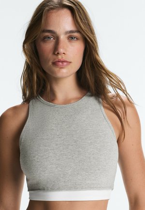 Jeune femme aux longs cheveux bruns portant un crop top gris sans manches sur un fond clair uni.