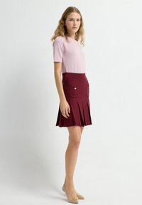 Korte mouw lichtroze top, gecombineerd met een bordeauxrode geplooide rok met zijzakken en gouden knoopaccenten. Lichtbeige schoenen.