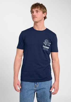 Mann mit kurzen Haaren, der ein marineblaues Hummel-T-Shirt mit kleinem weißem Motiv und hellblaue Jeans vor einem einfarbigen Hintergrund trägt.