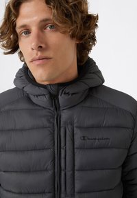 Czarna pikowana kurtka z kapturem, z zamkiem błyskawicznym i kieszenią na piersi. Materiał ma gładką, błyszczącą fakturę z pikowanymi detalami.