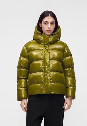 Une femme porte une doudoune matelassée vert olive brillante, avec une grande capuche et un pantalon noir, sur un fond clair uni.