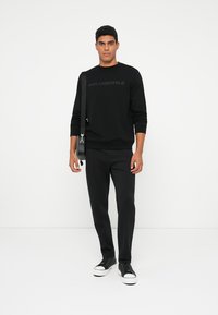 Felpa nera in cotone con logo in rilievo, abbinata a pantaloni neri e sneakers nere testurizzate. Modello che tiene una borsa a tracolla nera.