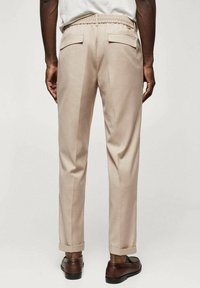 Beżowe spodnie o luźnym kroju, z elastycznym pasem, dwoma tylnymi kieszeniami i podwiniętymi mankietami, w połączeniu z brązowymi butami. Gładka tekstura materiału.