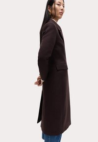 Długi, ciemnobrązowy płaszcz o dopasowanym kroju, z klapy kołnierza i pojedynczym zapięciem na guzik. Posiada boczne rozcięcia i przednie kieszenie.