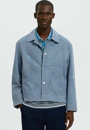 Jeune homme portant une veste en daim bleu clair, une chemise rayée et un pantalon foncé, debout devant un fond clair uni.
