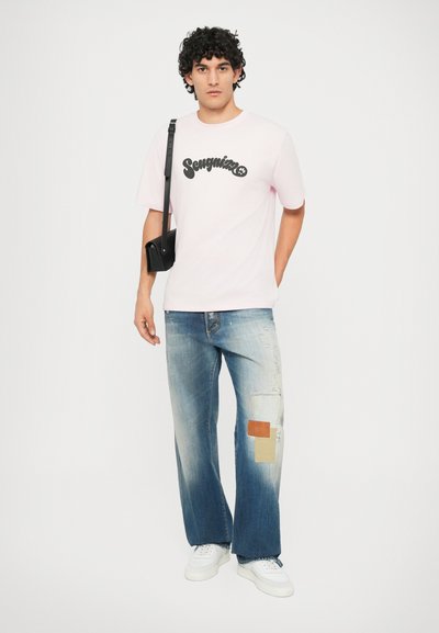 T-shirt rosa a maniche corte con grafica nera "Songmin", abbinato a jeans a gamba larga azzurri strappati con toppe e sneakers bianche.