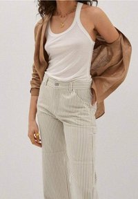 Bílý žebrovaný top bez rukávů pod hnědou lněnou košilí, doplněný kalhotami s vysokým pasem a širokými nohavicemi v světle zelených a bílých vertikálních pruzích.