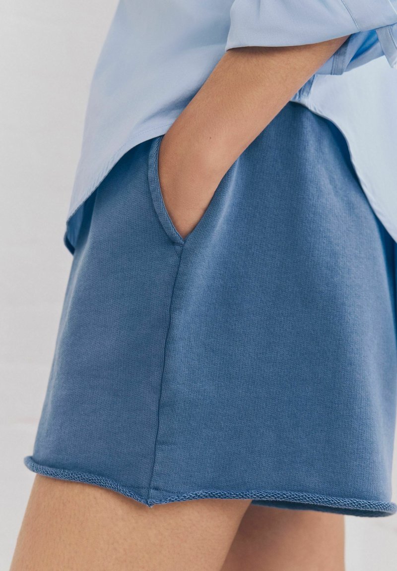 Pantaloni corti in cotone blu con una texture liscia, dotati di tasche laterali e orli cuciti, abbinati a una camicia azzurro chiaro visibile sopra.