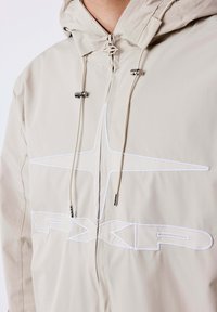 Giacca beige con cappuccio, zip frontale, cordini con estremità in metallo e stella ricamata bianca con logo PXLP sul petto.