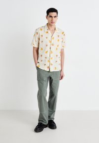 Camisa tropical de manga corta en color blanco roto con estampados de frutas coloridas, combinada con pantalones cargo verde oliva y zapatos negros.