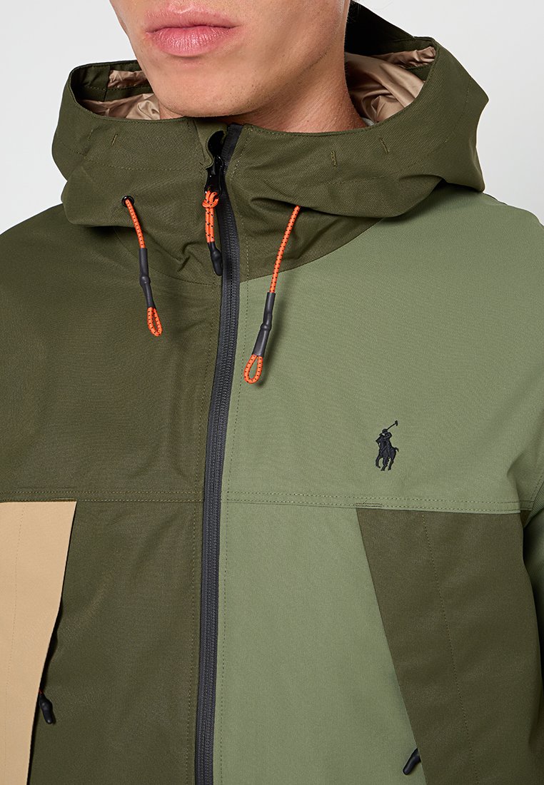 Giacca con cappuccio in verde oliva e design a blocchi di colore beige. Presenta una zip frontale, cordini regolabili e un piccolo logo nero. Tessuto testurizzato.