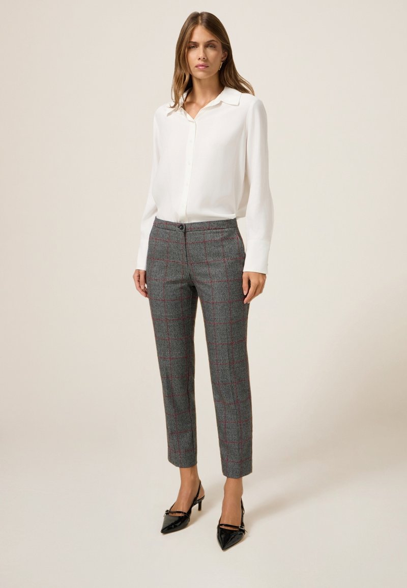 Camicia bianca con bottoni e maniche lunghe abbinata a pantaloni a quadri grigi con accenti rossi, completata da tacchi a punta neri.