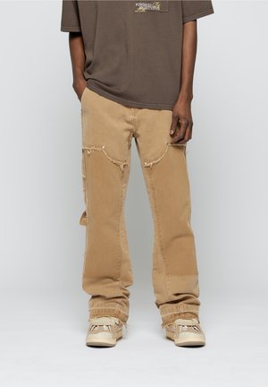 Personne portant un pantalon large beige usé, des baskets beiges avec des lacets tressés, et un t-shirt marron avec un texte jaune sur la poitrine.