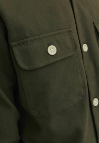 Olivově zelená košile na knoflíky, vyrobená z měkké látky, s dvěma kapsami na hrudi a bílými knoflíky a s pečlivým prošíváním podél okrajů.