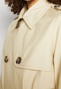 Gros plan sur un trench beige avec des boutons en écaille porté par une personne aux cheveux bouclés, mettant l’accent sur le col et la partie supérieure de la poitrine.