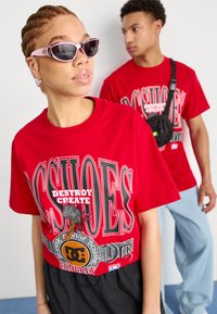 T-shirts graphiques rouges avec le texte "DÉTRUIRE CRÉER". Une personne porte des lunettes de soleil en forme de chat rose ; l'autre a un sac bandoulière noir.