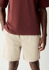 Chemise bordeaux à manches courtes avec une poche, associée à un short en coton beige. Les deux tissus ont une texture, une coupe décontractée et des bords ourlés.