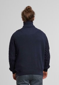 Maglione lavorato a maglia blu navy con colletto alto, caratterizzato da maniche a trecce testurizzate e schiena liscia. Inclusi polsini e orlo a coste.