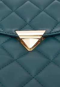 Teal kviltad läderhandväska med ett framträdande guld triangulärt spänne. Stickade detaljer förstärker textur och visuell som attraktionskraft.