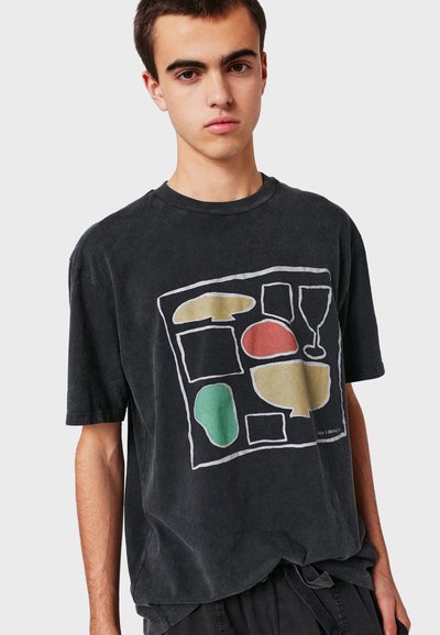 Un jeune homme portant un t-shirt noir oversize avec des formes colorées abstraites et des contours d'un verre sur le devant.