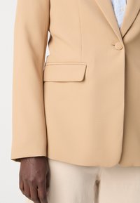 Blazer beige ajusté avec des épaules structurées, fermeture à un bouton et une poche latérale ; le tissu a une texture lisse et un léger éclat.