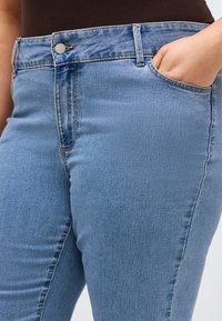 Άτομο που φοράει μπλε τζιν τζιν με το χέρι να στηρίζεται στην μπροστινή δεξιά τσέπη, μπροστά σε απλό φόντο.