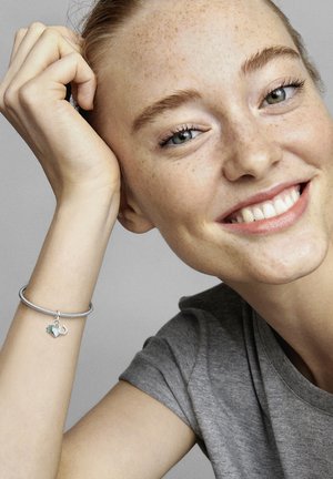Armband aus Sterlingsilber mit Charm und grünem Akzent. Das Modell trägt ein graues T-Shirt, lächelt und hat Sommersprossen sowie blaue Augen.