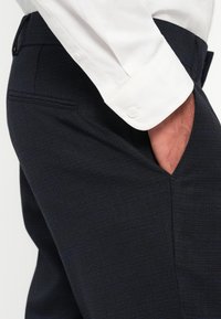 Pantalones oscuros de tela texturizada, con un corte ajustado y un ligero brillo, combinados con una camisa blanca de botones.