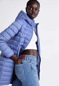 Casaco acolchoado azul claro com capuz, apresentando acolchoamento horizontal e um fecho frontal, combinado com jeans de lavagem média e uma blusa branca.