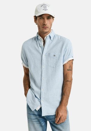 Lichtblauwe button-up shirt gemaakt van stof met een zachte textuur, met opgerolde mouwen en een borstzak. Draagt een witte pet.