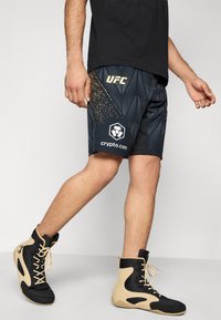 Μαύρα αθλητικά σορτς με χρυσό λογότυπο "UFC", υφή με σχέδιο φύλλων και σήμανση "crypto.com". Φοριούνται με μαύρα και μπεζ παπούτσια τύπου high-top.