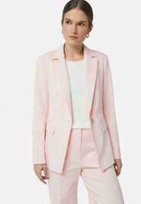 Blazer su misura rosa chiaro con revers a punta, bottone singolo e due tasche frontali. Abbinato a un top a coste bianco e pantaloni coordinati.