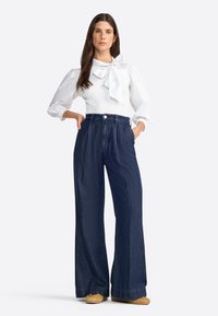 Blouse blanche avec un détail de nœud et des manches bouffantes, associée à un jean en denim bleu foncé à taille haute et jambes larges. Des chaussures marron complètent le look.