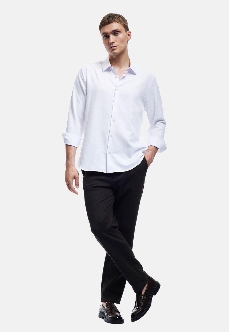 Witte langemouwen shirt met knoopsluiting, gecombineerd met zwarte broeken en glanzende zwarte schoenen. Glad materiaal en getailleerde pasvorm.